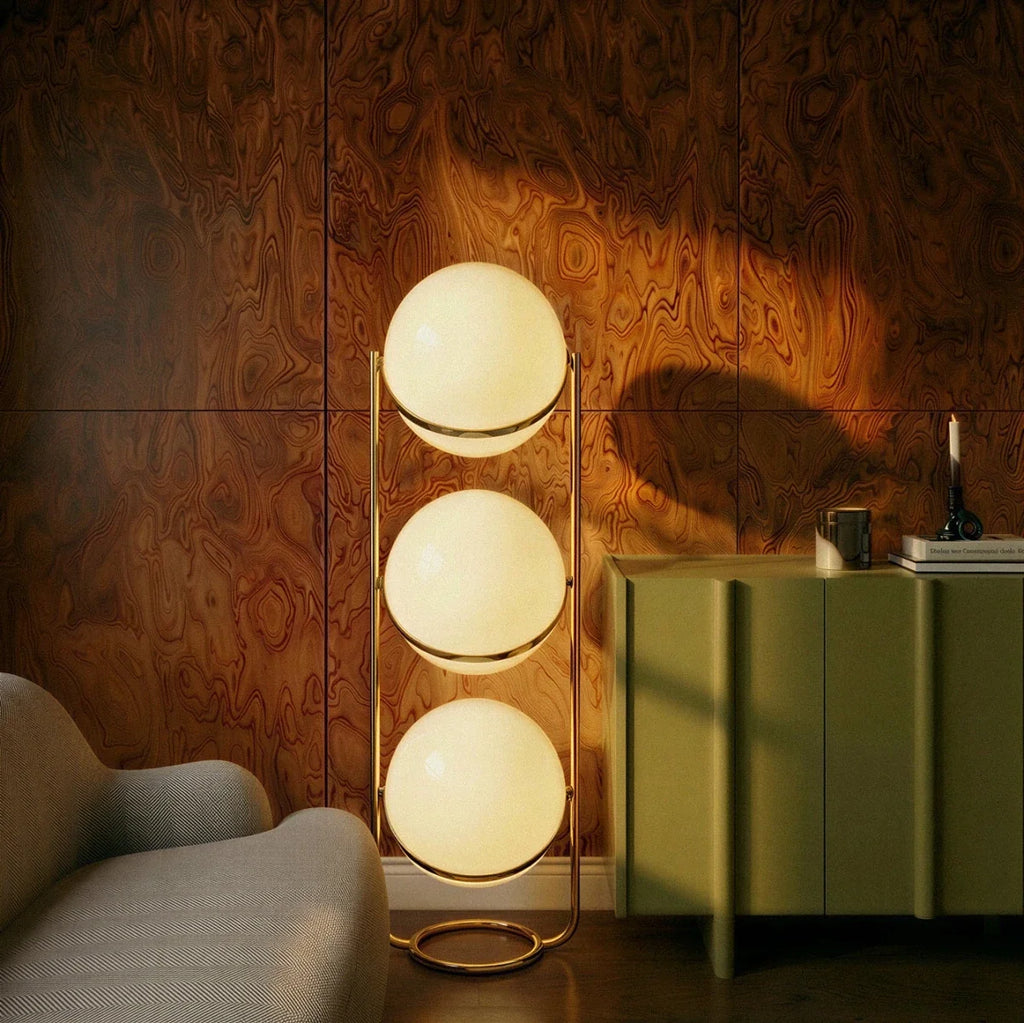 Clérachic three-globe sphere lamp in brass, lit beside an olive-green cabinet and throwing its shadow across a burl-wood panelled wall.