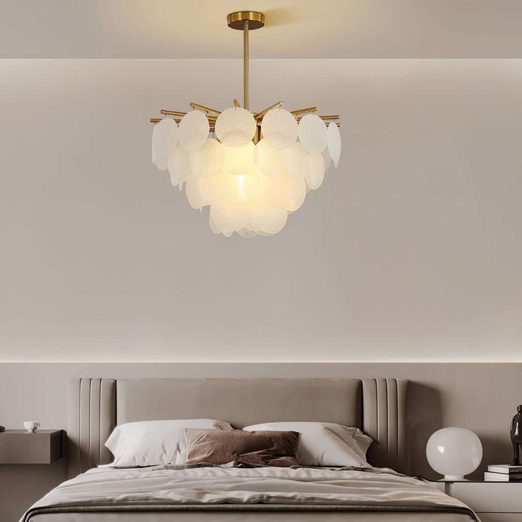 Small Clérachic disc chandelier lit above a bed with a taupe headboard, with a round white bedside lamp at the right.