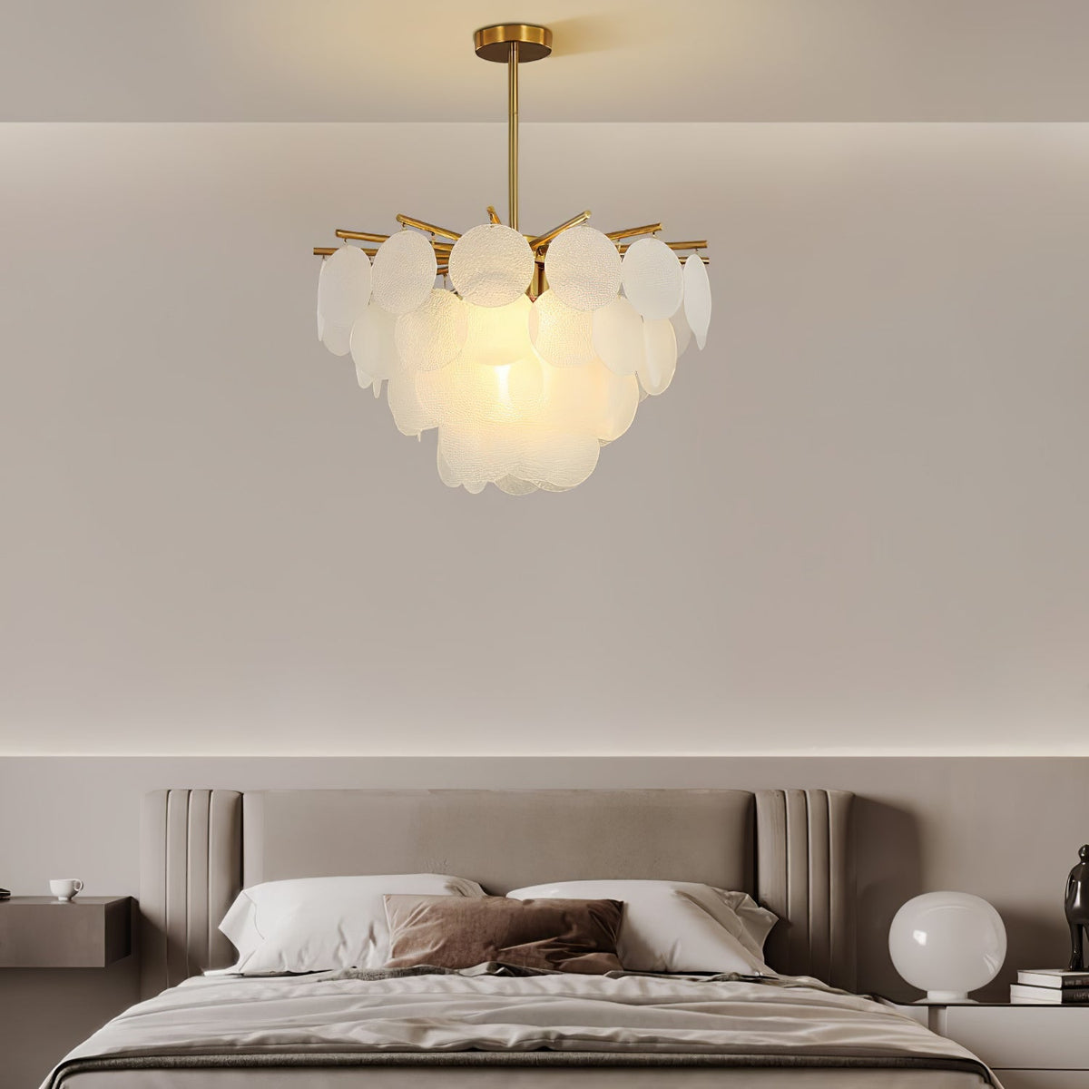 Small Clérachic disc chandelier lit above a bed with a taupe headboard, with a round white bedside lamp at the right.