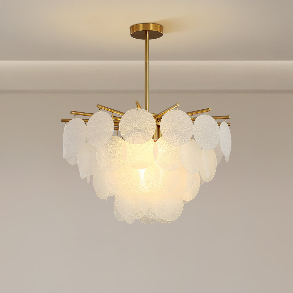 The upper tier of round textured discs and brass arms on this Clérachic chandelier, lit warmly, in a plain pale room.