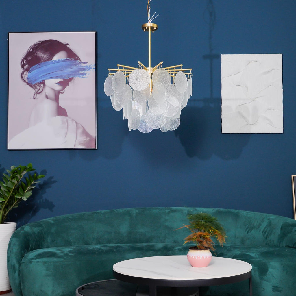 A Clérachic disc chandelier small in the frame against a deep blue wall, above a green velvet curved sofa and a round table.