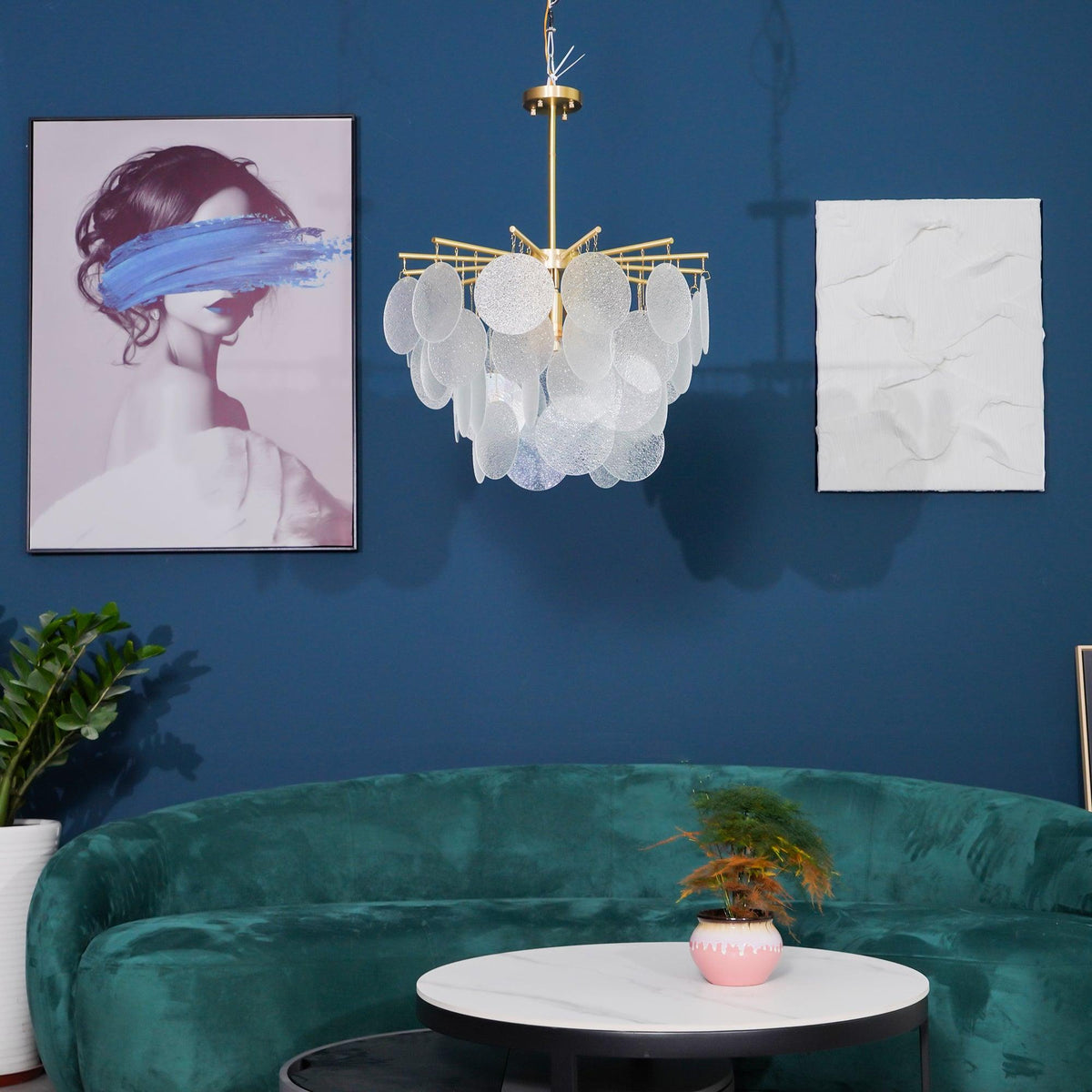A Clérachic disc chandelier small in the frame against a deep blue wall, above a green velvet curved sofa and a round table.