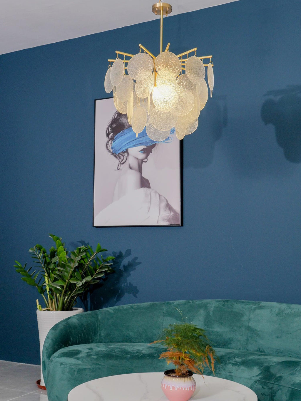 Close crop of this Clérachic chandelier, lit amber-white, hanging before a blue wall and a framed portrait with a blue brushstroke.