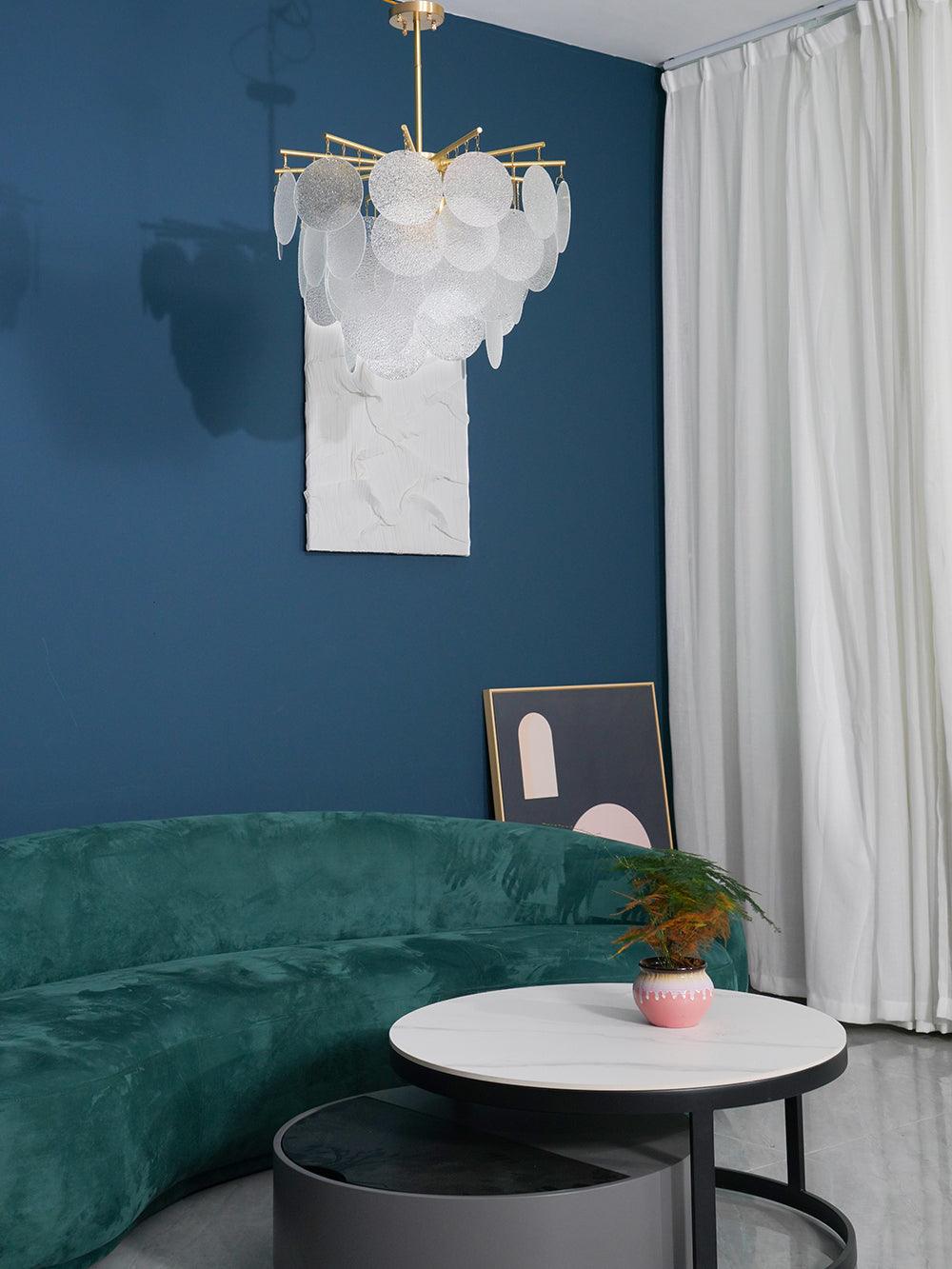 A Clérachic disc chandelier, unlit and pale, hangs in the corner of a blue-walled room with a curtain and a green velvet sofa.