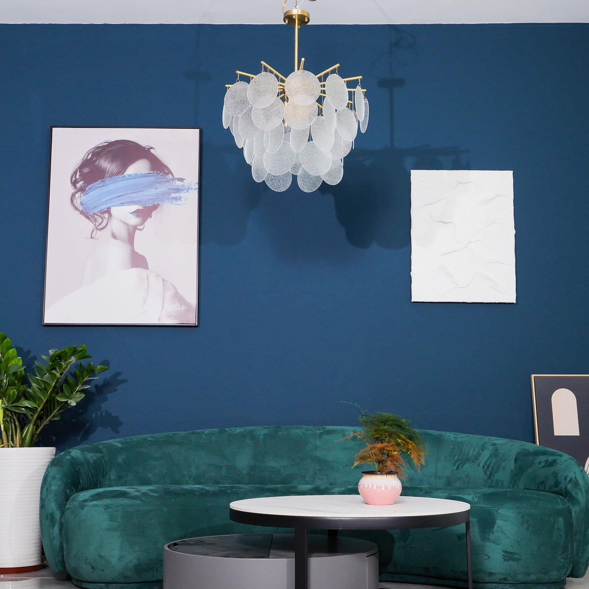Unlit Clérachic chandelier of frosted discs on a blue wall, with a framed portrait, a white textured panel and a green sofa below.