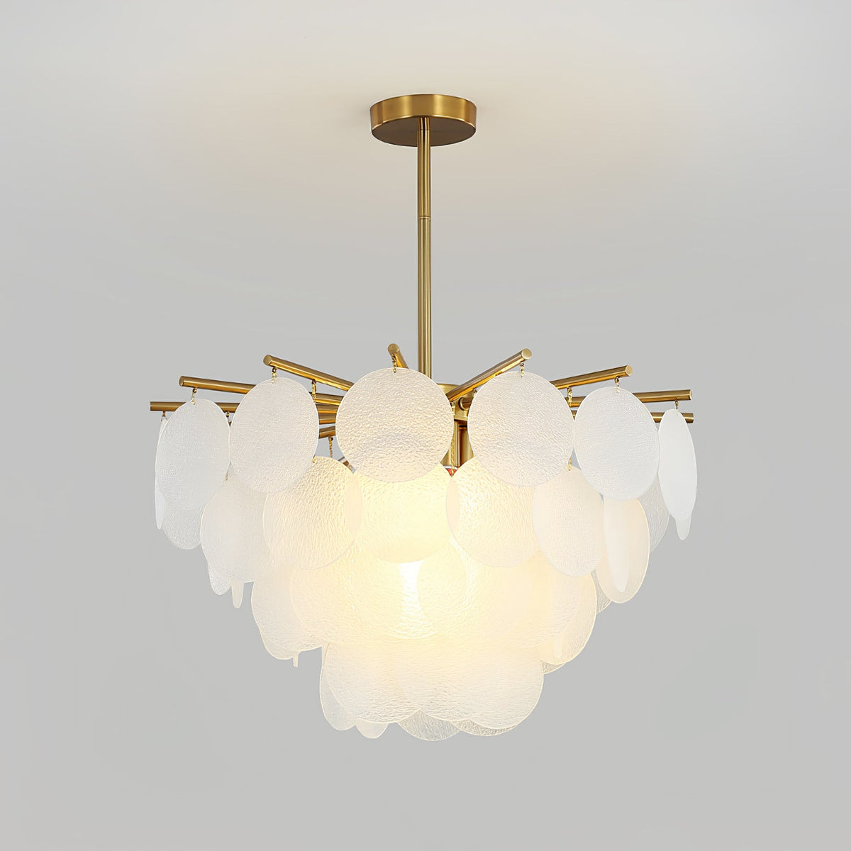 A Clérachic chandelier lit warmly in front of a plain grey backdrop, its frosted discs layered above a brass rod and canopy.