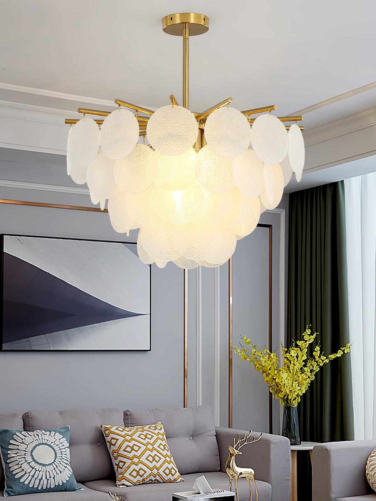 Lit Clérachic chandelier in a living room with a white cornice, patterned cushions on a grey sofa and a vase of yellow flowers.