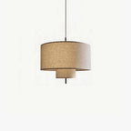 Clérachic pendant with a wide woven linen drum shade and a smaller tier beneath it, hung from a black cord on a plain white backdrop.