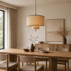 Two-tier linen Clérachic pendant glowing above a long oak dining table, with a dried-branch vase and a framed landscape behind.