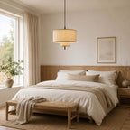 Tiered Clérachic pendant glowing over a bed with white and oatmeal linen, a woven bench at its foot and trees outside the window.