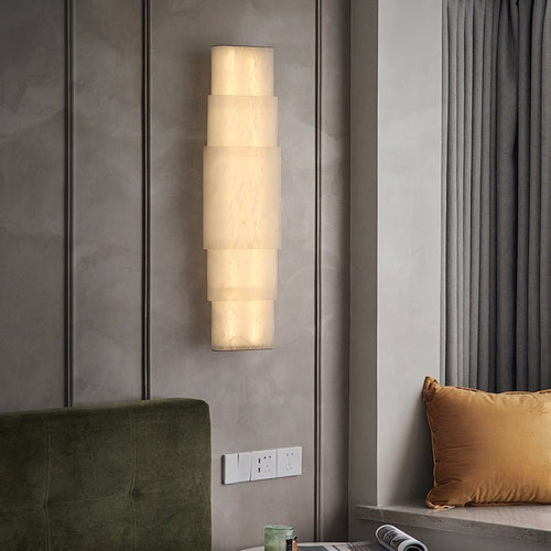 Clérachic tiered sconce lit beside a green velvet headboard, warm glow washing the grey wall.