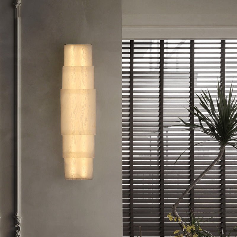 The Clérachic tiered sconce glowing on a grey wall next to venetian blinds and a potted palm.