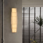 The Clérachic tiered sconce glowing on a grey wall next to venetian blinds and a potted palm.