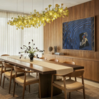 Clérachic's chandelier in its linear form, glowing gold above a long dining table beside sheer curtains.