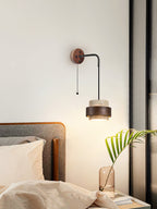 Lit above a bedside, a Clérachic wall light with a pull chain throws a warm glow beside a bed and a palm leaf in a glass vase.