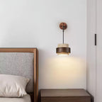 This Clérachic wall light shines a soft pool of warm light down a white wall beside a leather-framed bed and a walnut nightstand.