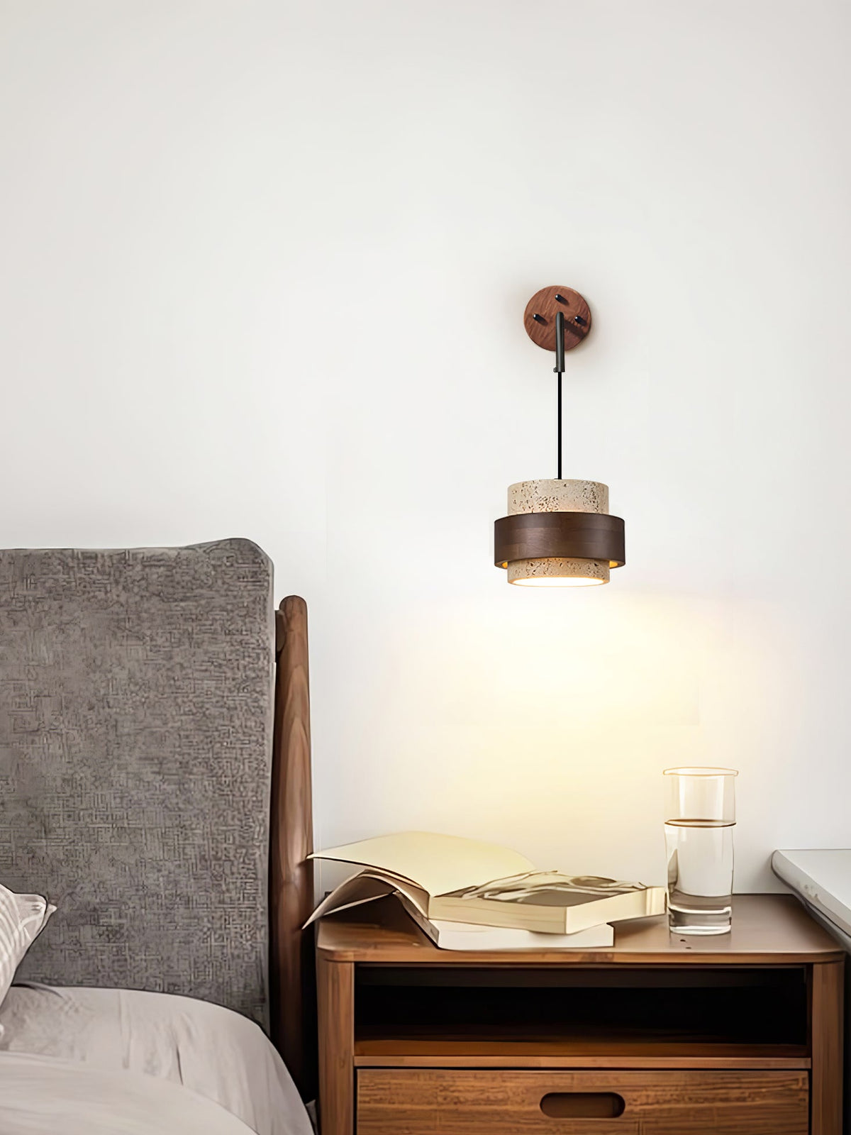 A Clérachic wall light lit above a wooden nightstand with an open book and a glass of water, next to a grey upholstered headboard.