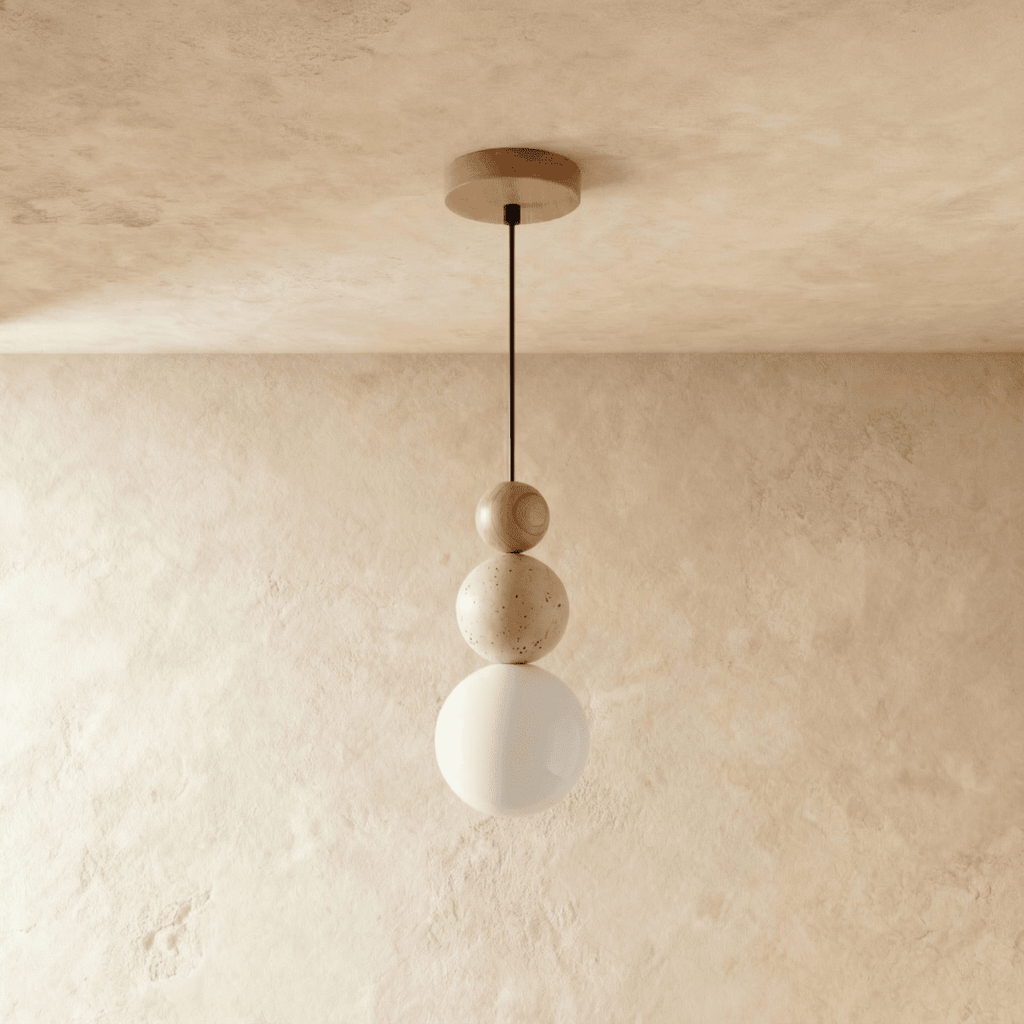 Clérachic pendant of a wood bead, a travertine bead and a white glass globe on a slim black cord, hung against limewash.