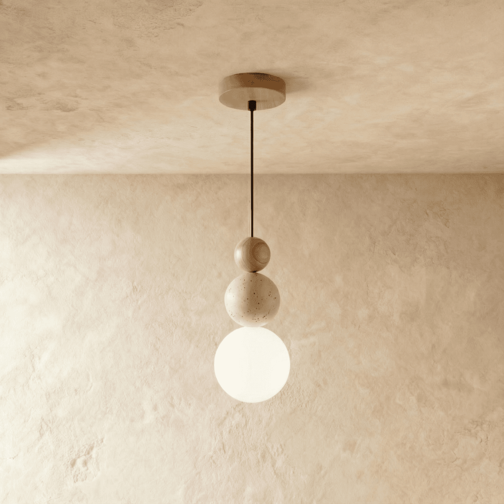 Clérachic stacked pendant with wood, speckled travertine and a lit opal white globe, on a black cord against a plaster wall.