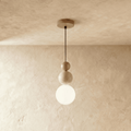 Clérachic stacked pendant with wood, speckled travertine and a lit opal white globe, on a black cord against a plaster wall.