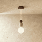 Clérachic stacked pendant with a dark walnut bead, travertine bead and white glass globe on a cord, against a limewashed wall.