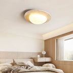 Flush-mounted Clérachic ceiling light glowing above a bedroom with a pale linen bed, a wooden wall panel and a window with sheer curtains.