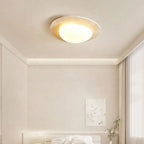 A Clérachic travertine-rimmed ceiling light in a calm cream bedroom with a concealed cove light, a small framed print and tall curtains.