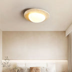 A Clérachic ceiling light above a cream bedroom wall, with a headboard, cushions and a leafy branch in the lower corner.
