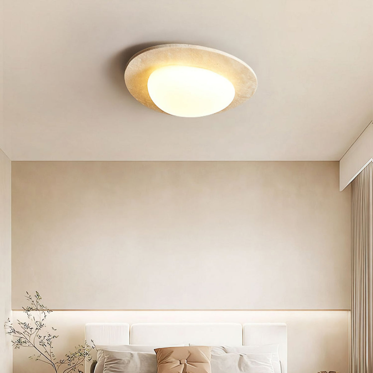 A Clérachic ceiling light above a cream bedroom wall, with a headboard, cushions and a leafy branch in the lower corner.