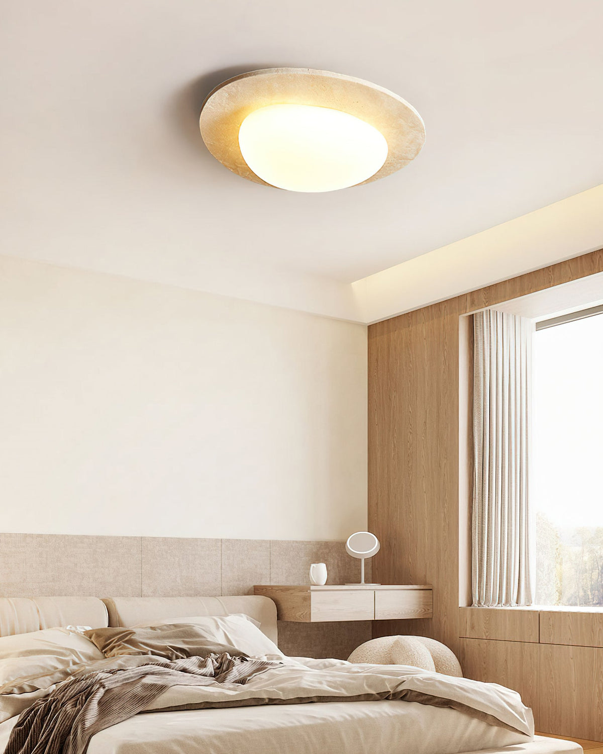 Clérachic travertine ceiling light seen from a low angle in a bedroom, with a wooden wall, a bedside shelf and a window with sheer curtains.