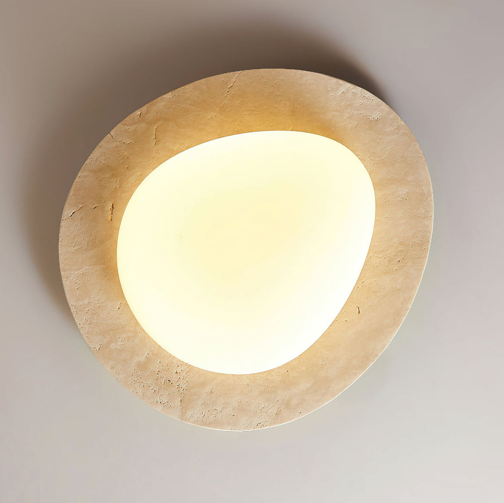 Looking up at this Clérachic ceiling light on a grey ceiling, with its uneven pale travertine rim and a warm-lit rounded glass shade.
