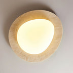 Looking up at this Clérachic ceiling light on a grey ceiling, with its uneven pale travertine rim and a warm-lit rounded glass shade.