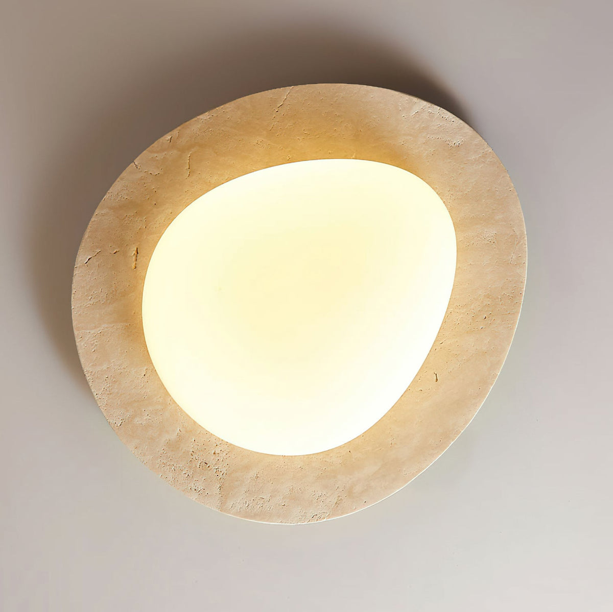 Looking up at this Clérachic ceiling light on a grey ceiling, with its uneven pale travertine rim and a warm-lit rounded glass shade.