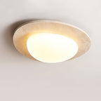 Lit Clérachic flush ceiling light from the side, its pebble-shaped glass shade glowing warm inside a pale stone rim.