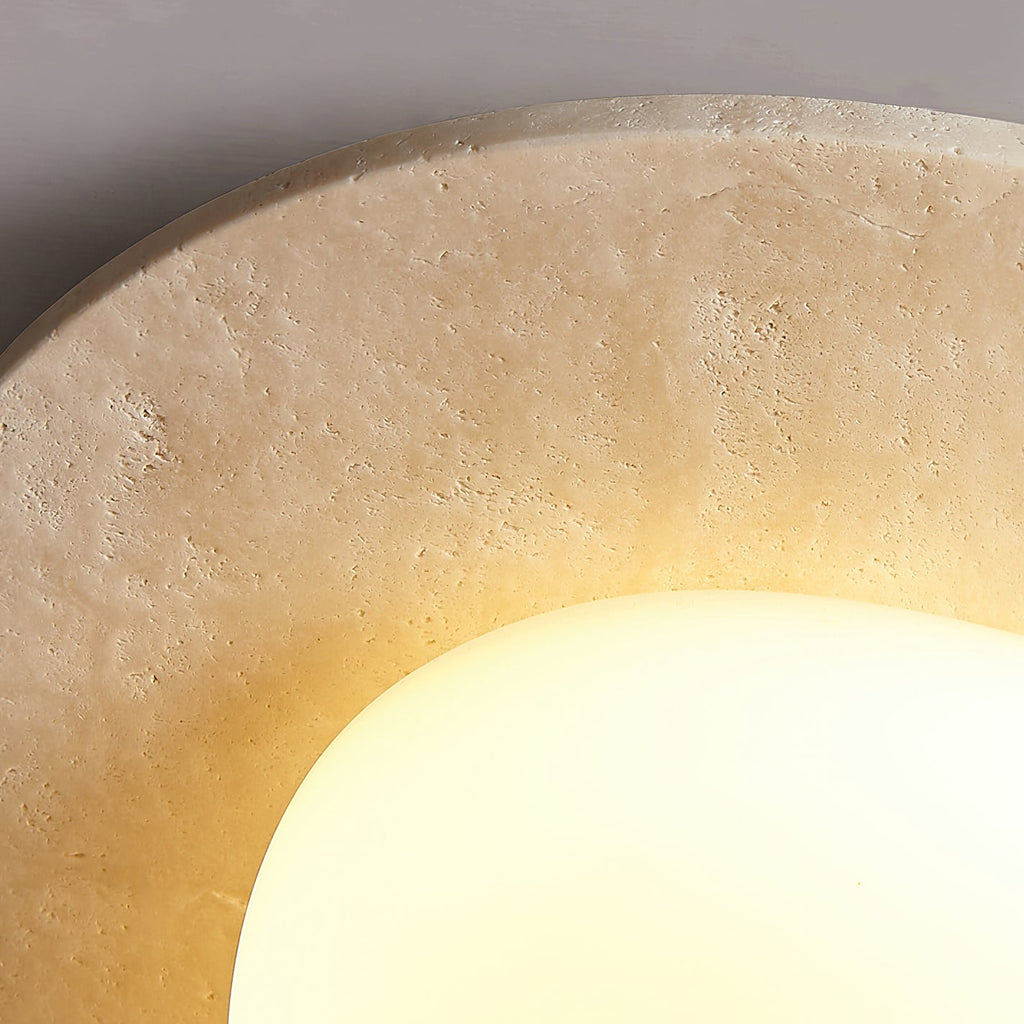Close view of the pitted, natural texture of the travertine rim on this Clérachic ceiling light beside the lit glass.