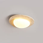 Lit Clérachic ceiling light photographed at a slant, with the glass shade offset inside its stone-look rim against a grey ceiling.