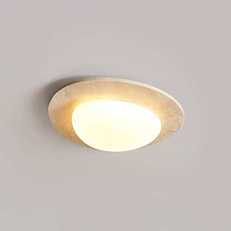 Lit Clérachic ceiling light photographed at a slant, with the glass shade offset inside its stone-look rim against a grey ceiling.