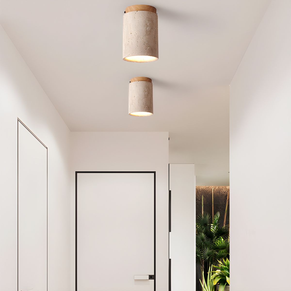 Two Clérachic travertine ceiling lights lit in a white hallway, one behind the other above a slim black-framed door.