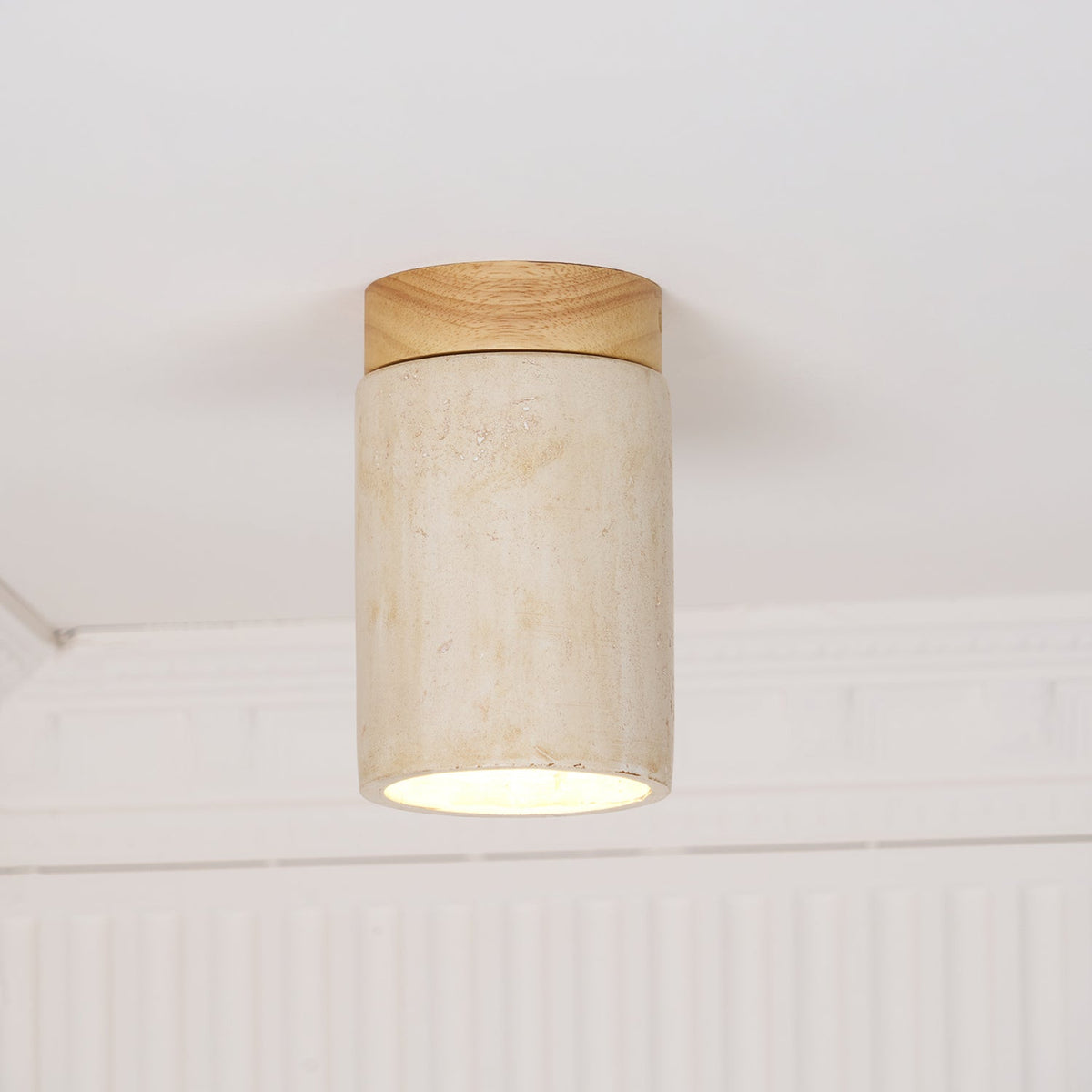 Front view of a Clérachic ceiling light, a cream travertine cylinder under a wood cap, with the glow showing at the rim.