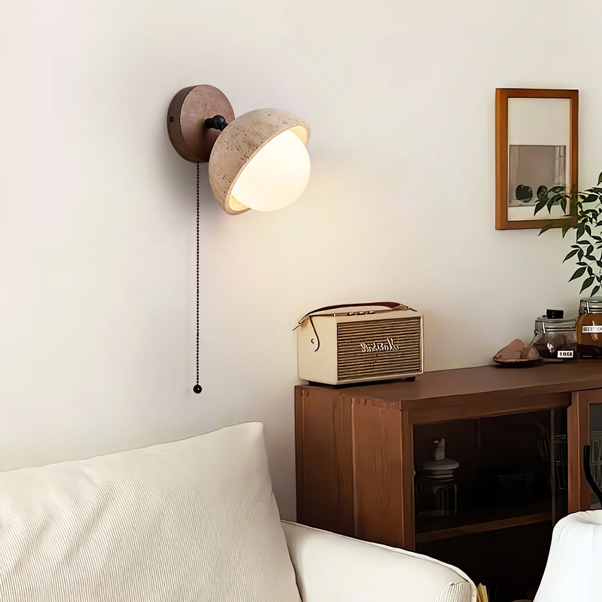 Pull-chain travertine wall light from Clérachic glowing beside a walnut cabinet with a retro radio, and a cream sofa below.