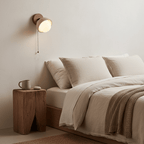 Clérachic wall light with a travertine shade and pull chain above a linen-dressed bed and a chunky wooden stump bedside table.