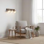 Triple-dome travertine wall light from Clérachic lit above a grey armchair and a woven rug in a bright corner by a window.