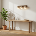 This Clérachic three-dome wall light is lit over a pale wooden console table, with a leafy plant in a terracotta pot beside it.