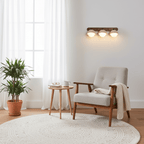 Clérachic three-dome wall light glowing on a white wall, near a wooden armchair, a round side table and a large indoor plant.