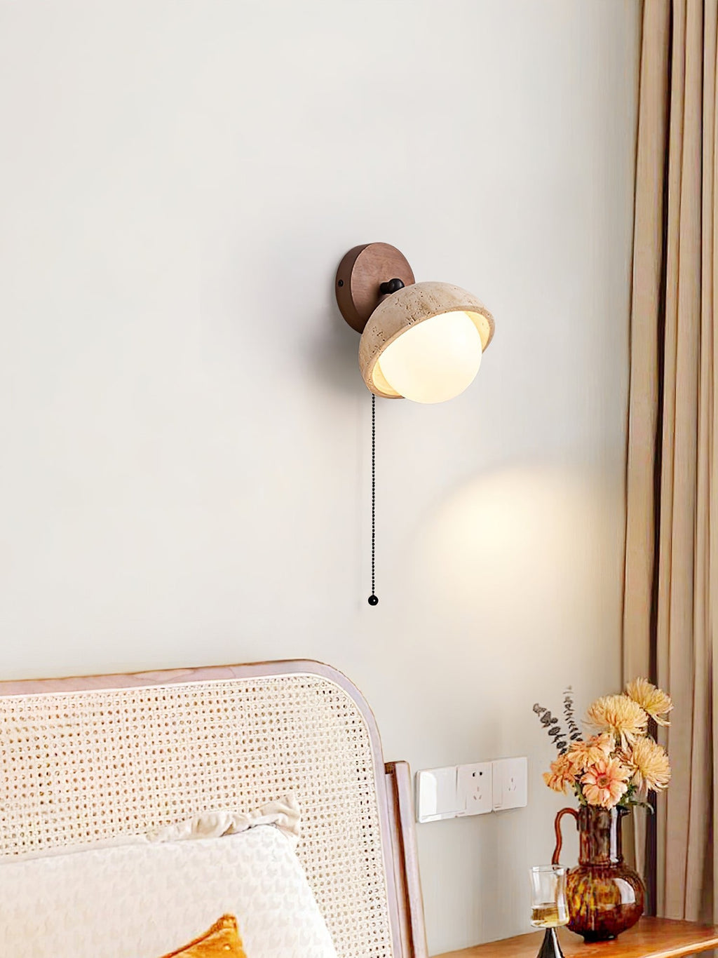 Single pull-chain Clérachic wall light with a travertine dome and a walnut round backplate, above a cane headboard and a window curtain.