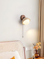 Single pull-chain Clérachic wall light with a travertine dome and a walnut round backplate, above a cane headboard and a window curtain.