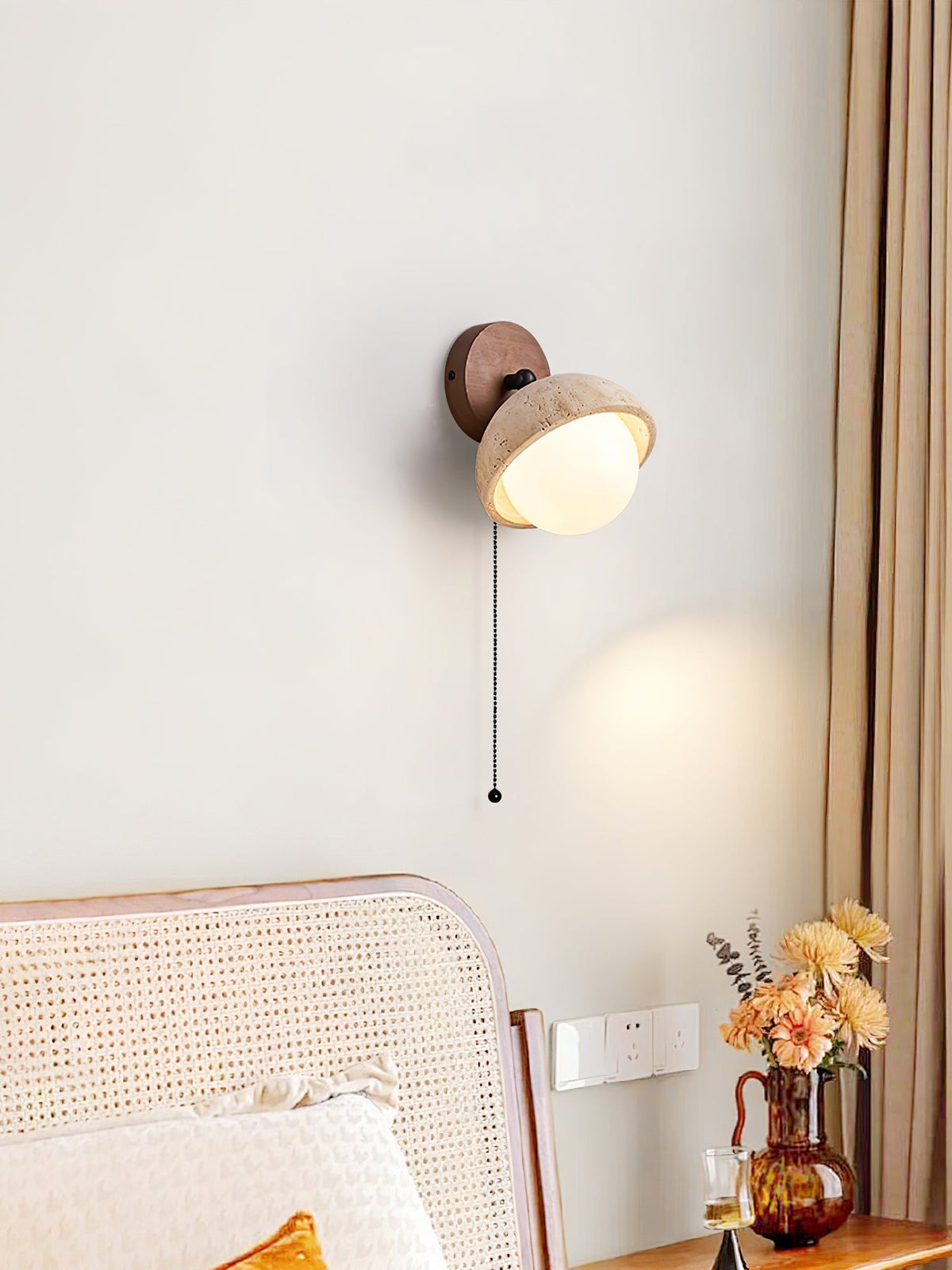 Single pull-chain Clérachic wall light with a travertine dome and a walnut round backplate, above a cane headboard and a window curtain.