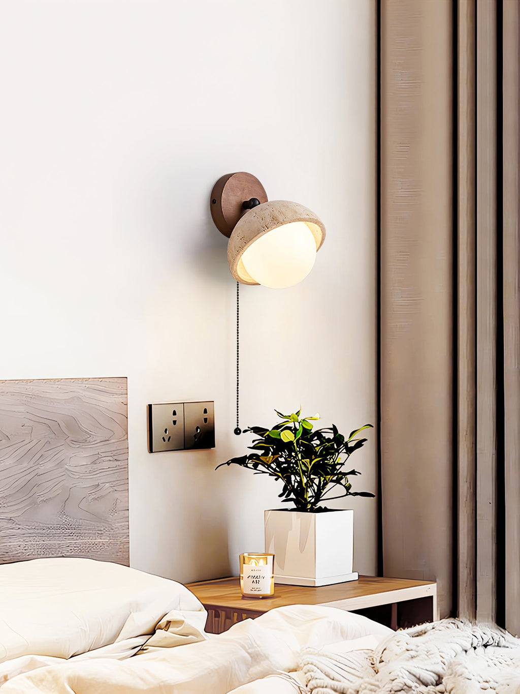 Travertine dome wall light from Clérachic with a pull chain, above a bedside table with a plant in a white pot and a lit candle.