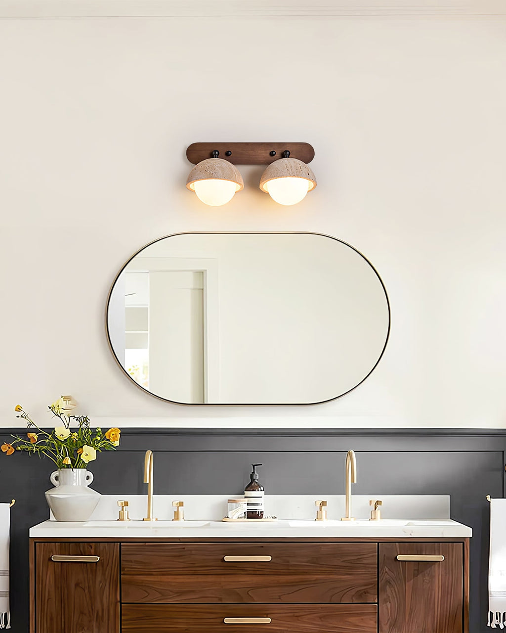 Two-dome Clérachic wall light lit above an oval black-framed mirror, with a walnut vanity and brass taps in a bathroom.