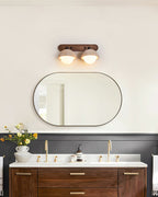 Two-dome Clérachic wall light lit above an oval black-framed mirror, with a walnut vanity and brass taps in a bathroom.